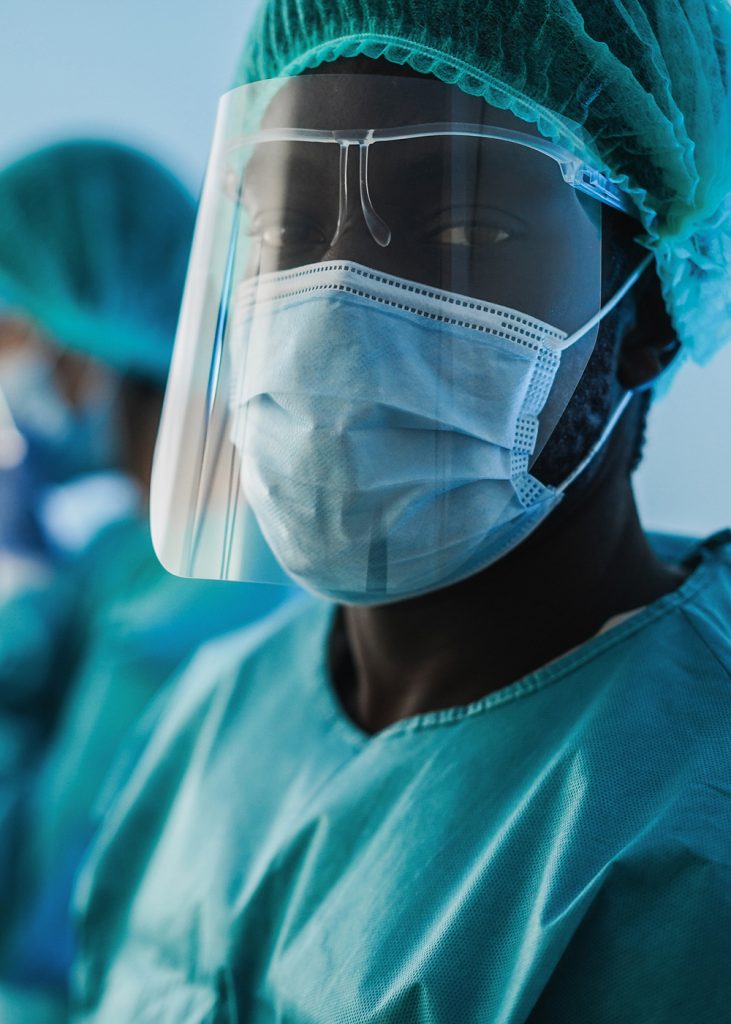 A black male doctor wearing teal scrubs, a surgical cap, a fact mask, and clear face shield, looking to camera.